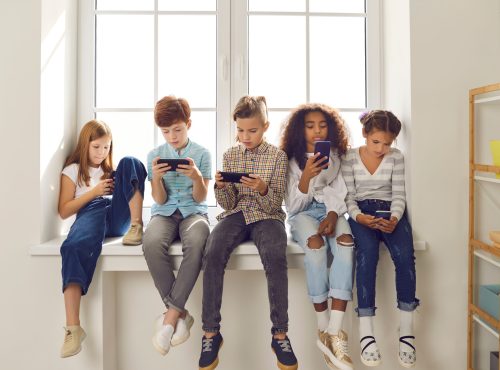 Group of children of different nationalities sit in a row on the windowsill and with lack of emotion play online games or read social networks on mobile phones. Technologies that spoil childhood.